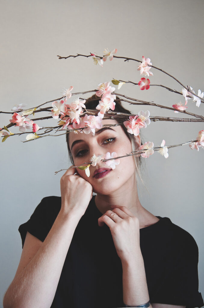 Darian Rojas poses gracefully with delicate cherry blossom branches intertwined around her face set against a muted background.
